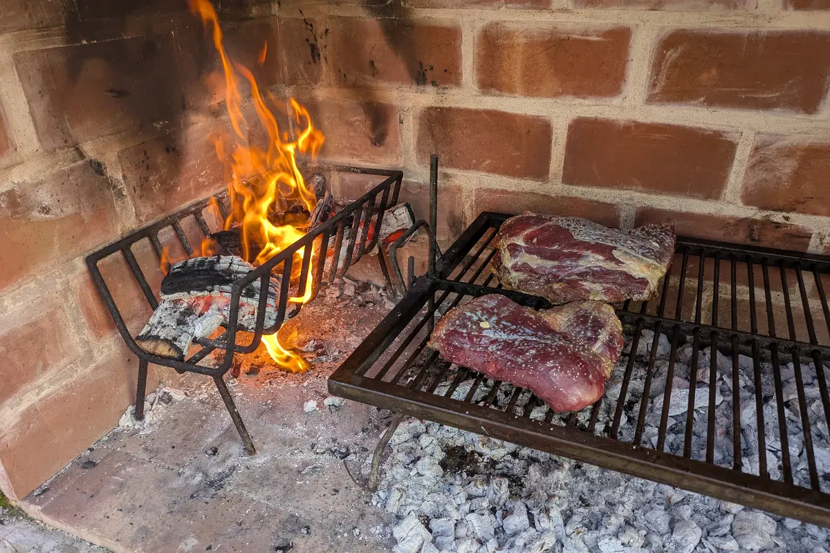 Two cuts of raw beef with coarse salt on an iron grill over embers in a brick hearth, with a basket of burning logs beside it